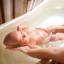 Small cute big-eyed beautiful baby bathes in warm water in the hands of a caring mother. The concept of baby care and child care